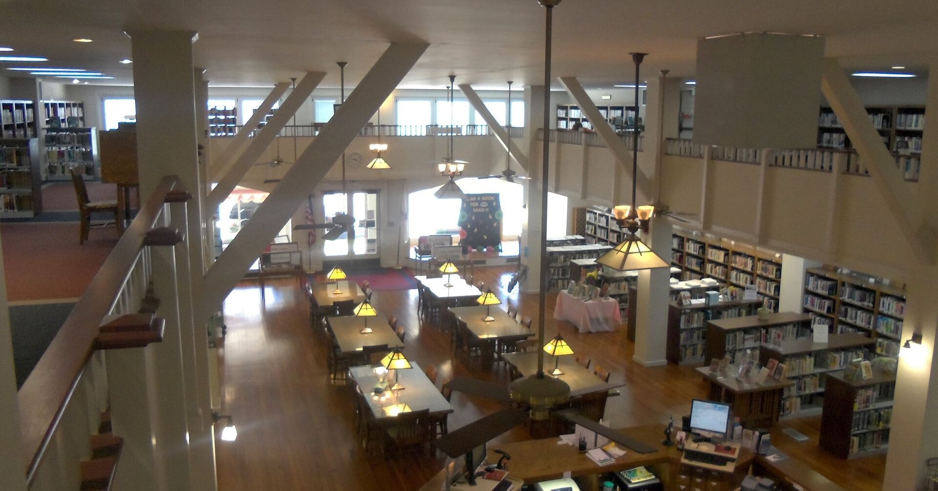 A spacious, two-story library with bookshelves lining the walls, wooden tables and chairs with lamps, computers on desks, and large windows allowing natural light to fill the room.