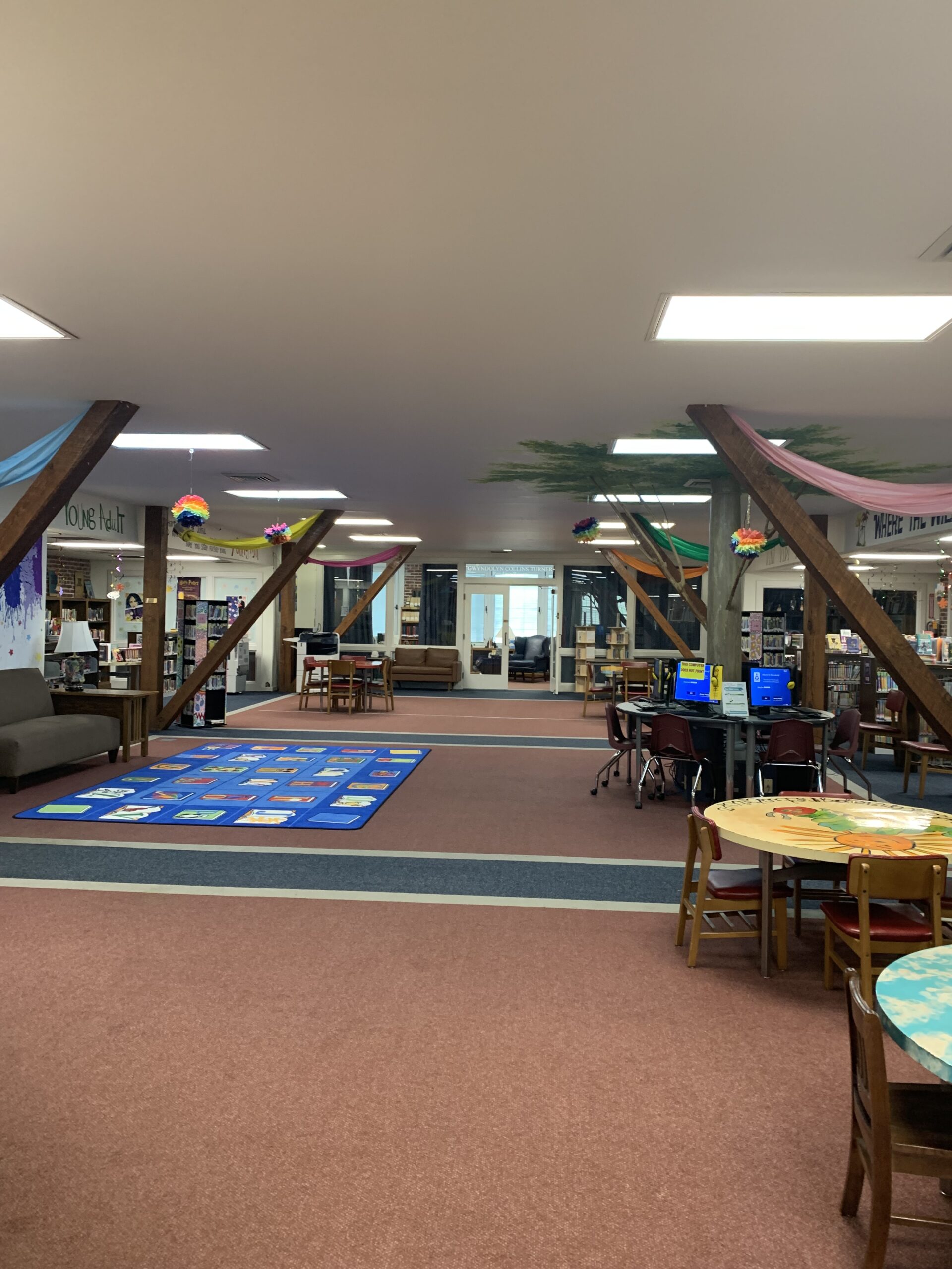 A spacious library with wooden beams, bookshelves, a colorful rug in the center, tables and chairs, computer stations, and hanging decorations from the ceiling. The atmosphere appears welcoming and organized.