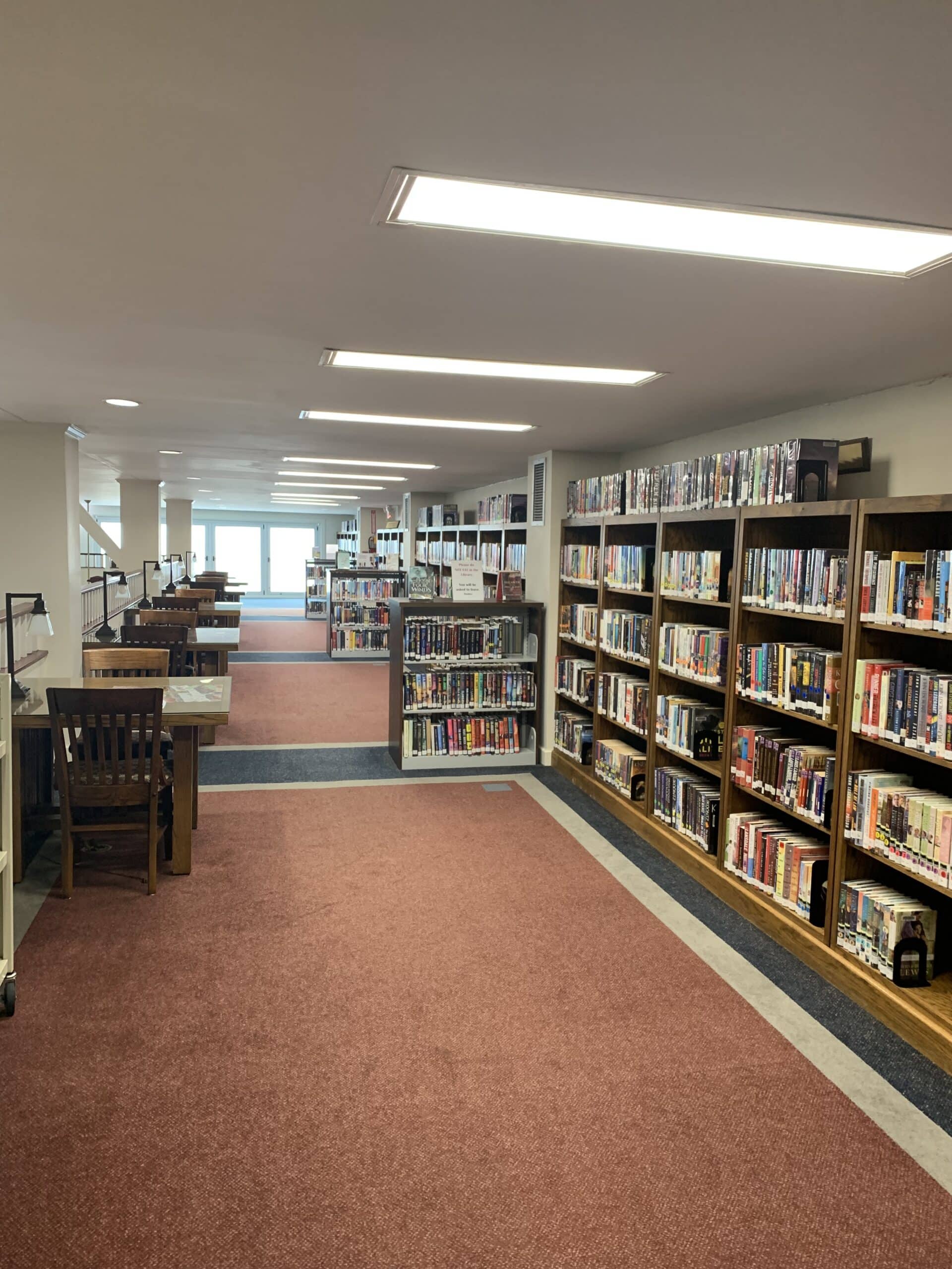 A quiet library interior with rows of bookshelves filled with books on the right, wooden study tables and chairs on the left, and large windows in the background letting in natural light.